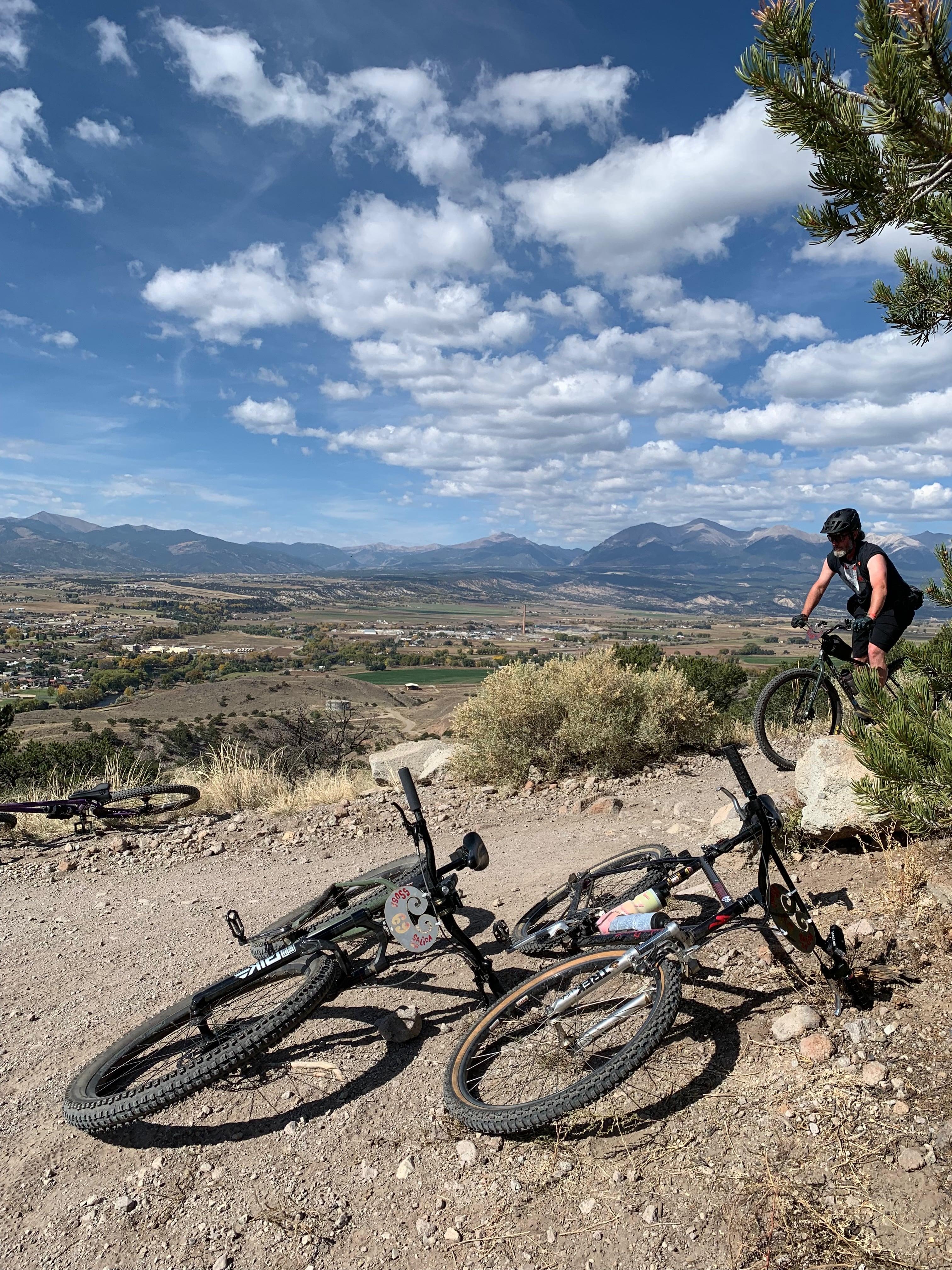 Rider looking out over the Salida valley from the trail, two bikes dropped in the foreground under a dramatic cloudy sky