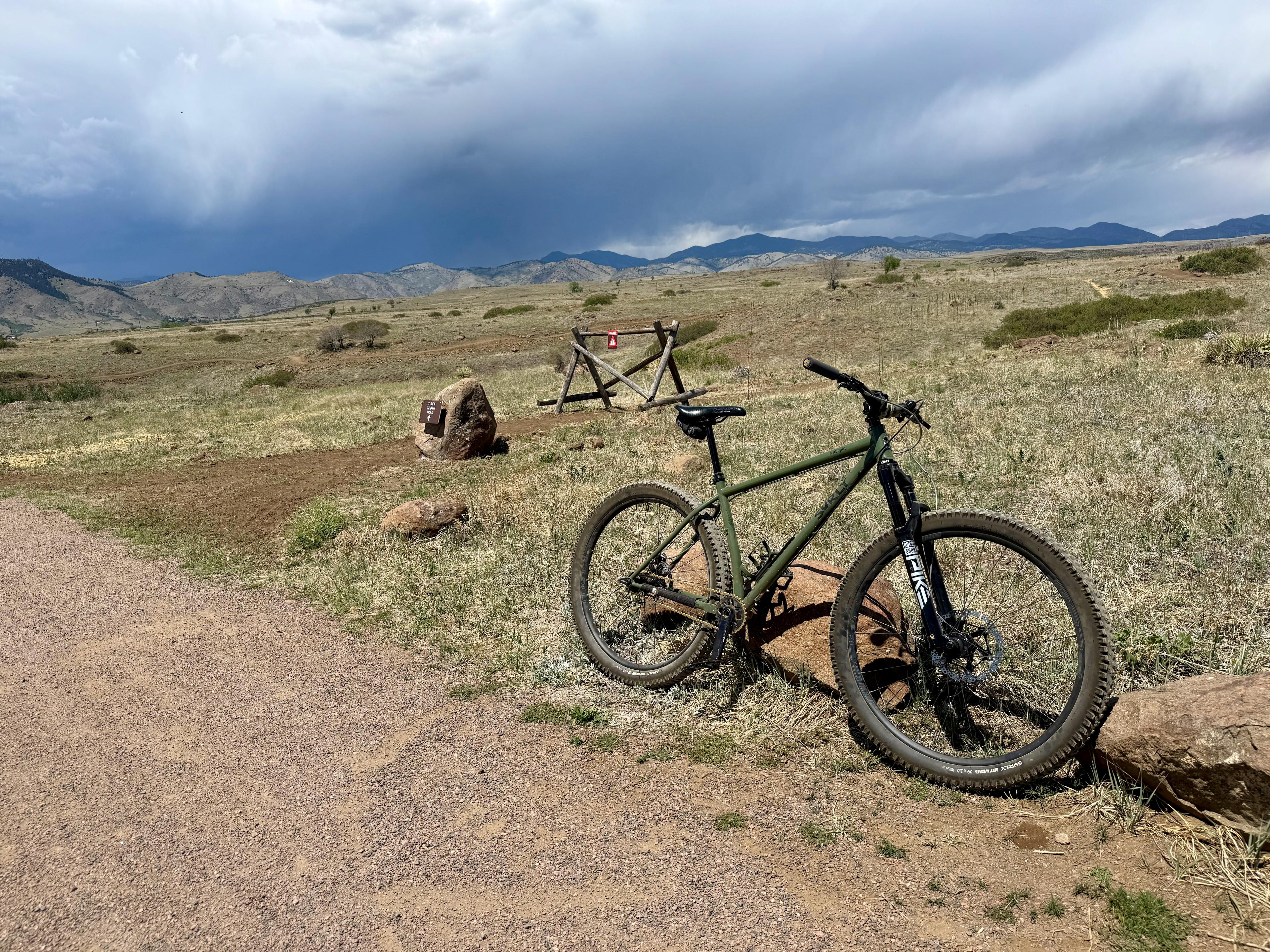 Surly Krampus single-speed mountain bike leaning on a rock beside a dirt trail, with storm clouds over the foothills behind it