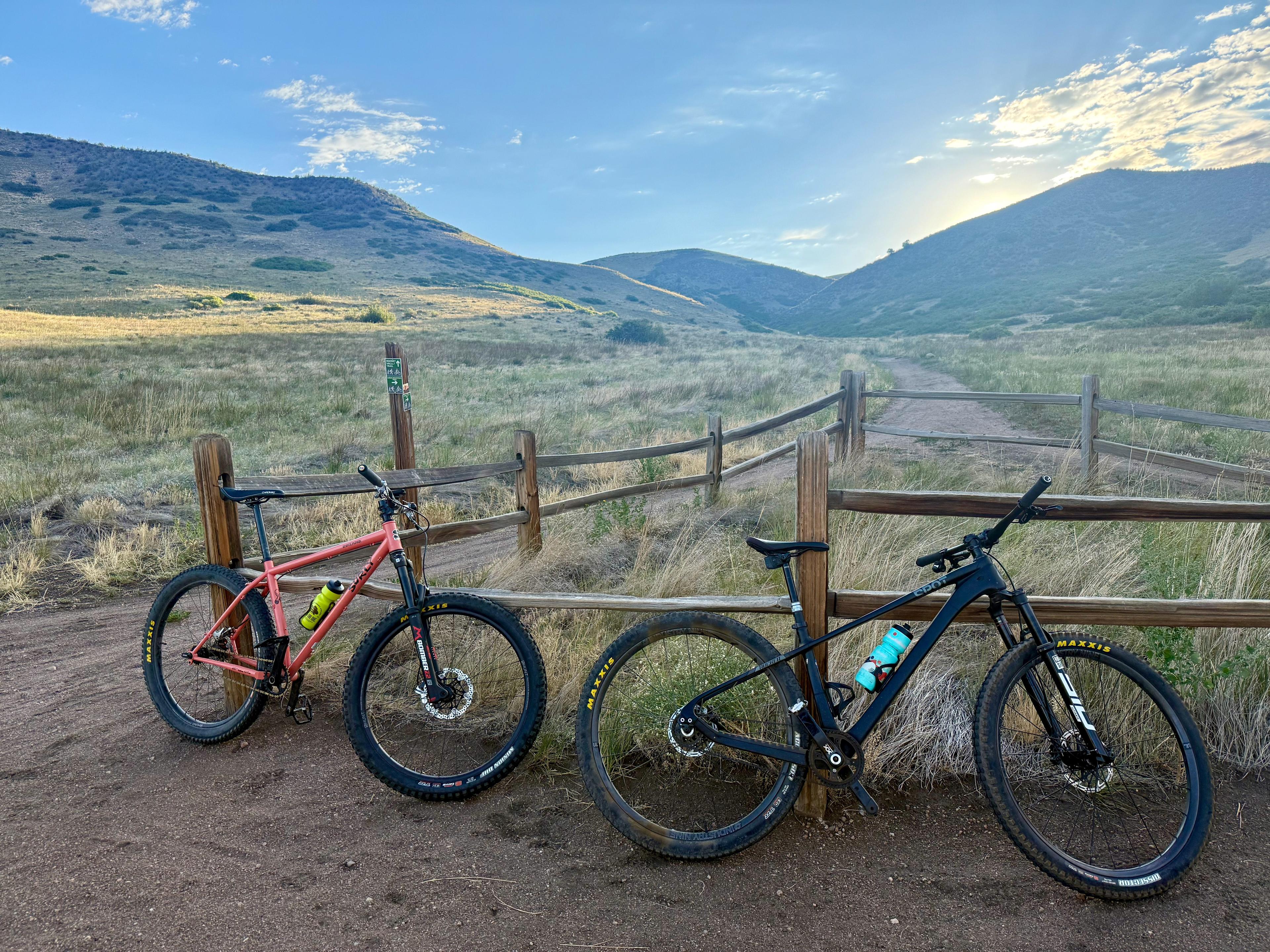 A salmon Surly Karate Monkey and a black Spot belt-drive bike leaning on a split-rail fence at a trailhead, a grassy valley opening toward low morning light