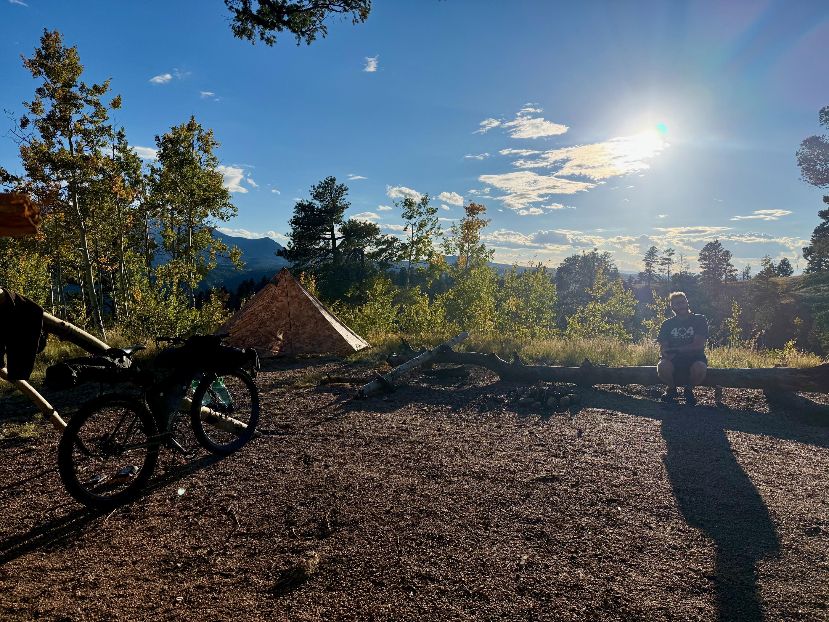 Blake sitting on a fallen aspen log at golden hour next to a pyramid tent at the dispersed camp, mountains silhouetted behind, loaded Krampus leaned against a tree in the foreground