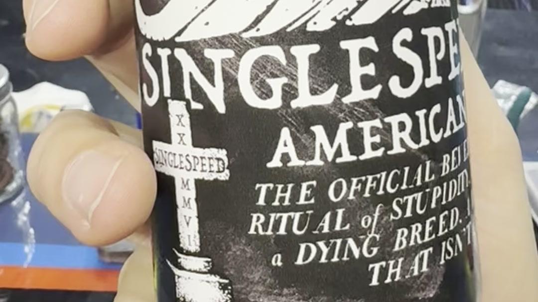 A hand turning a black can of Singlespeed American Pale Ale, the white label text reading the official beverage of our ancient ritual of stupidity and a world class event that isn't classy at all