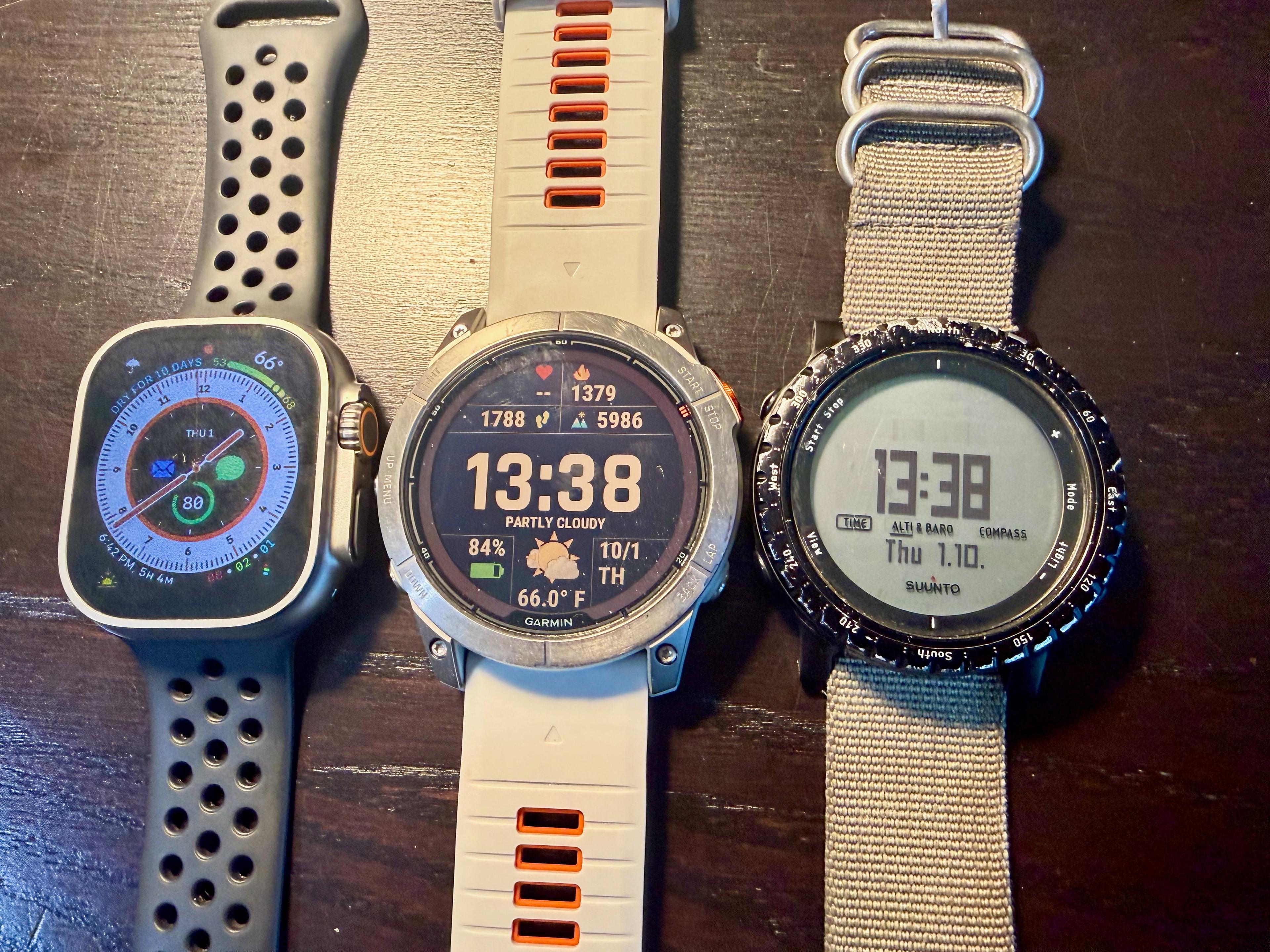 Apple Watch Ultra, Garmin Fenix 7X Pro Sapphire Solar, and Suunto Core lined up side by side on a wood table, all showing the same time