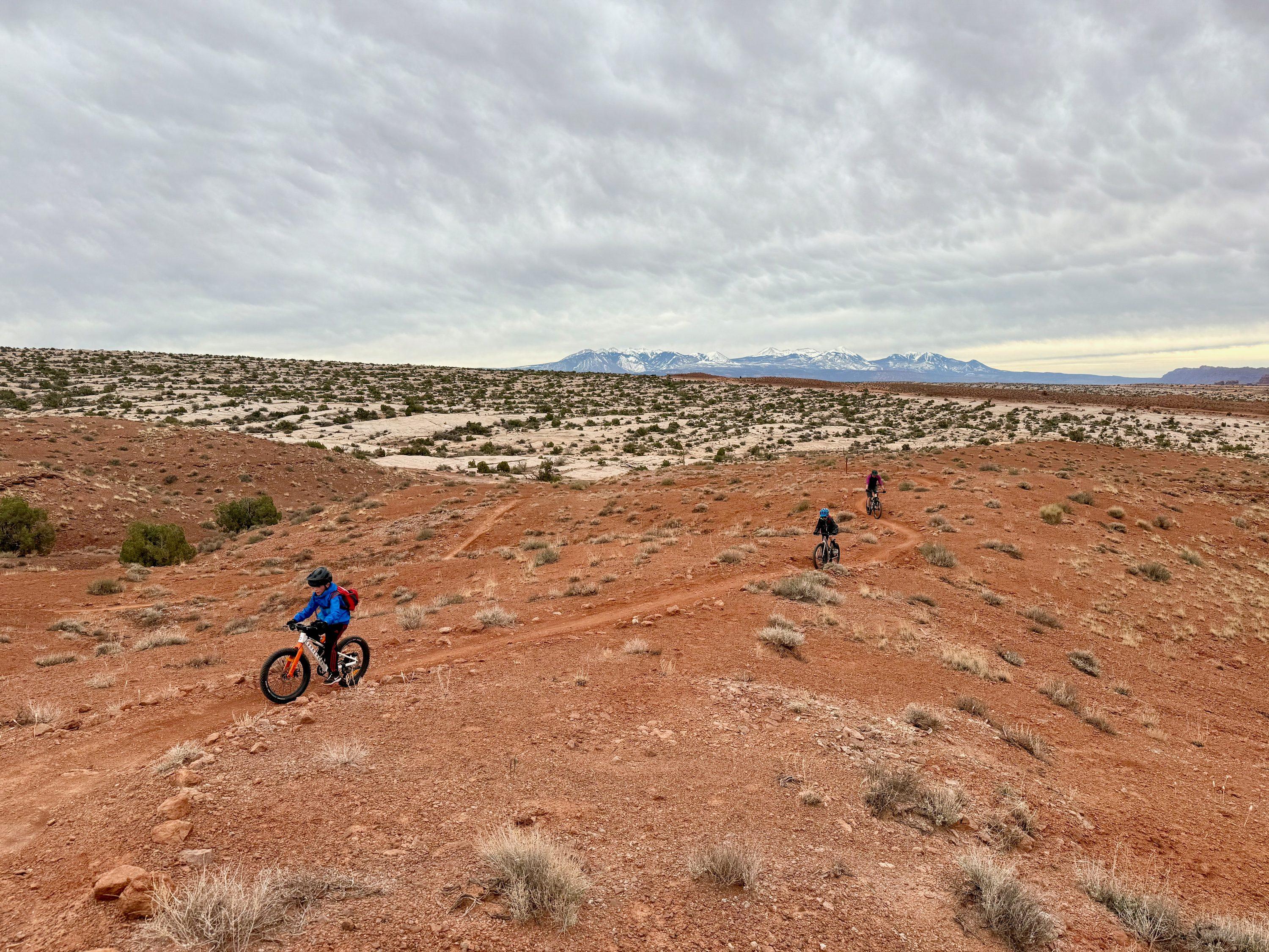 Three mountain bikers strung out along a red dirt singletrack across the Moab desert, the snow-capped La Sal Mountains on the horizon under a heavy gray sky